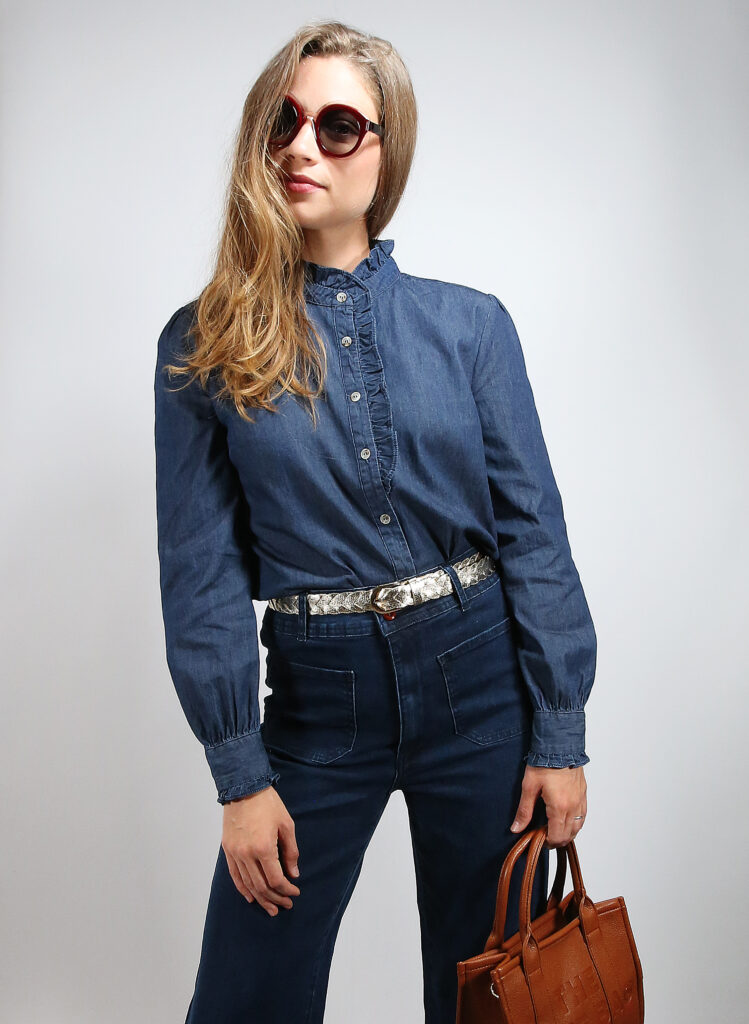 Jeune femme prise en photo dans un studio, avec une tenue mode : joli chemisier à volants, jean flare avec poche, ceinture dorée et sac à main façon Totebag de grande marque.