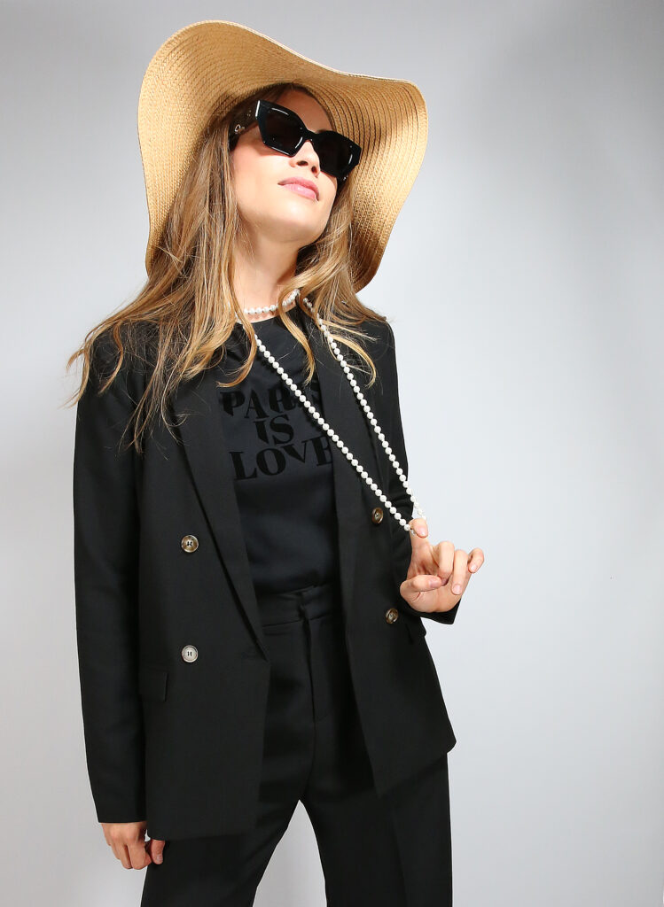 Jeune femme prise en photo dans un studio, avec une tenue total look noir mode : un tailleur veste pantalon, un t-shirt avec inscription "Paris is Love", un collier de perle inspiration Chanel, une paire de lunettes de soleil ultra glamour et un chapeau vogue qui termine la silhouette.